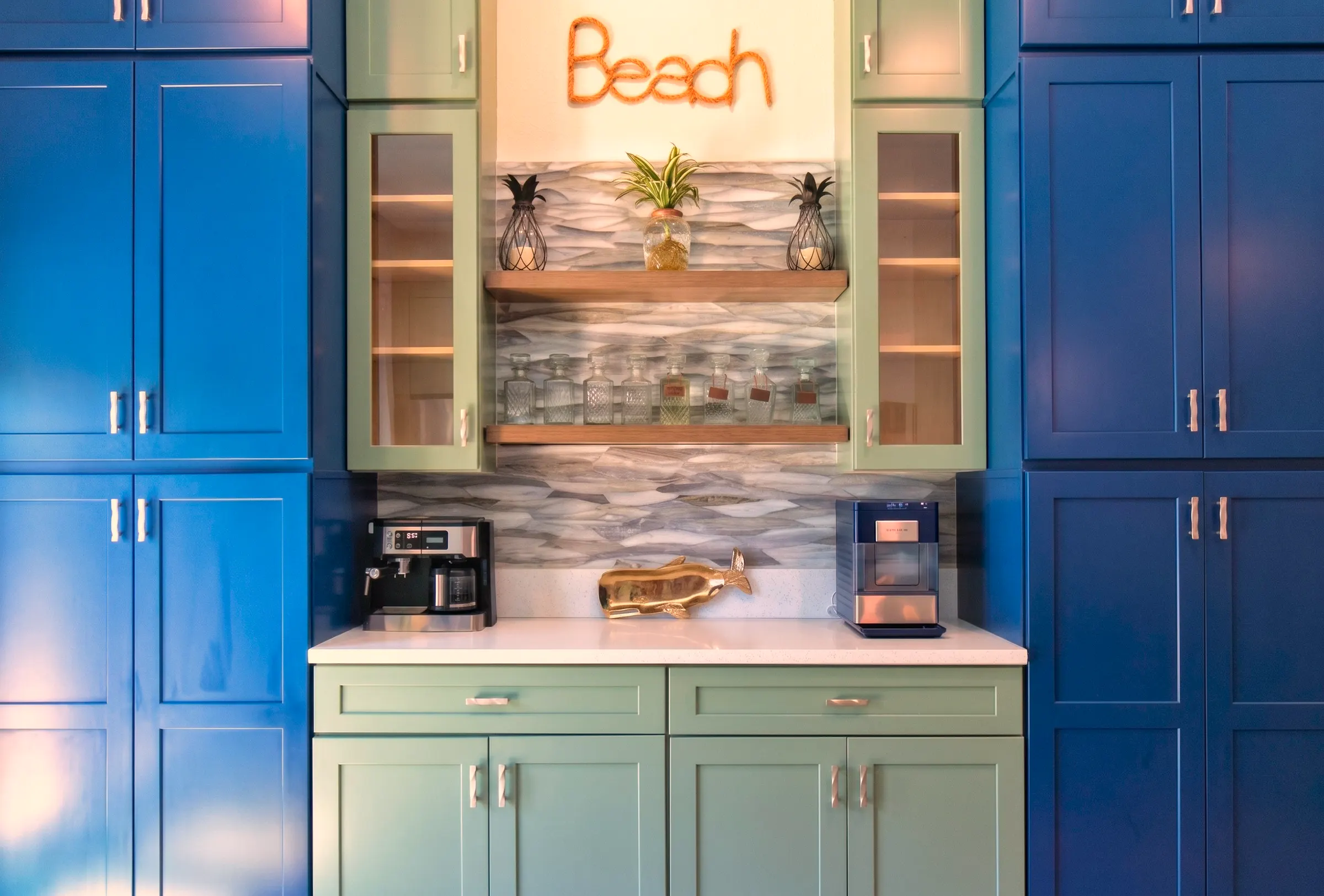 Beverage bar in sage green cabinetry set between floor-to-ceiling navy pantry towers, with a glass wave-tile backsplash and a floating wood shelf of glass decanters