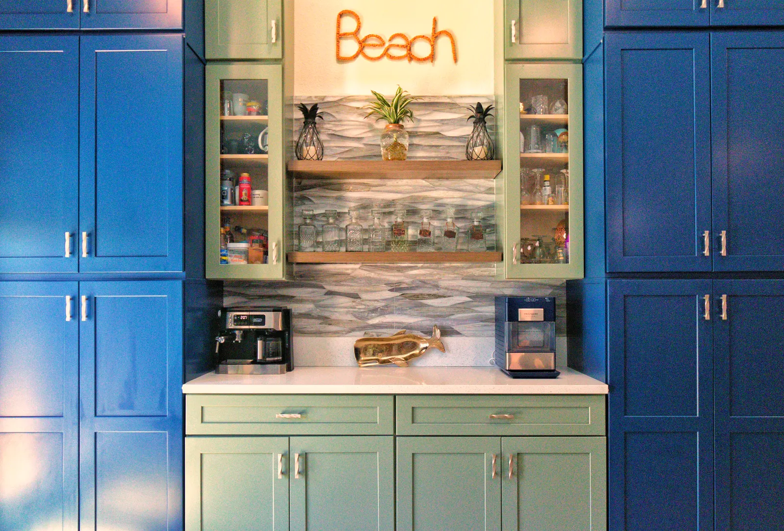 Wider view of the sage beverage bar and navy pantry towers, with a wire script sign reading Beach above the glass shelving