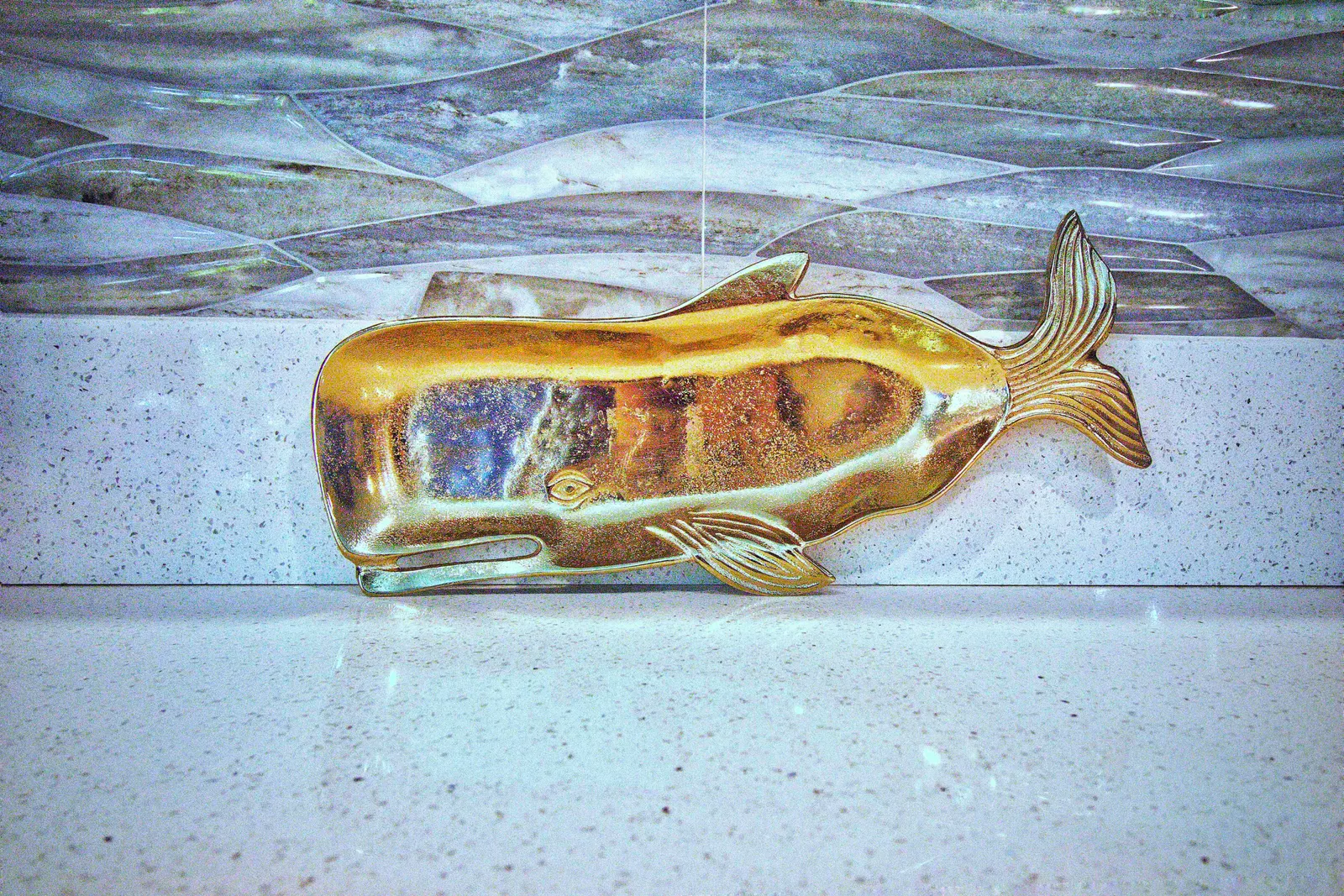 Brass whale sculpture resting on a white speckled quartz counter against the gray glass wave-tile backsplash
