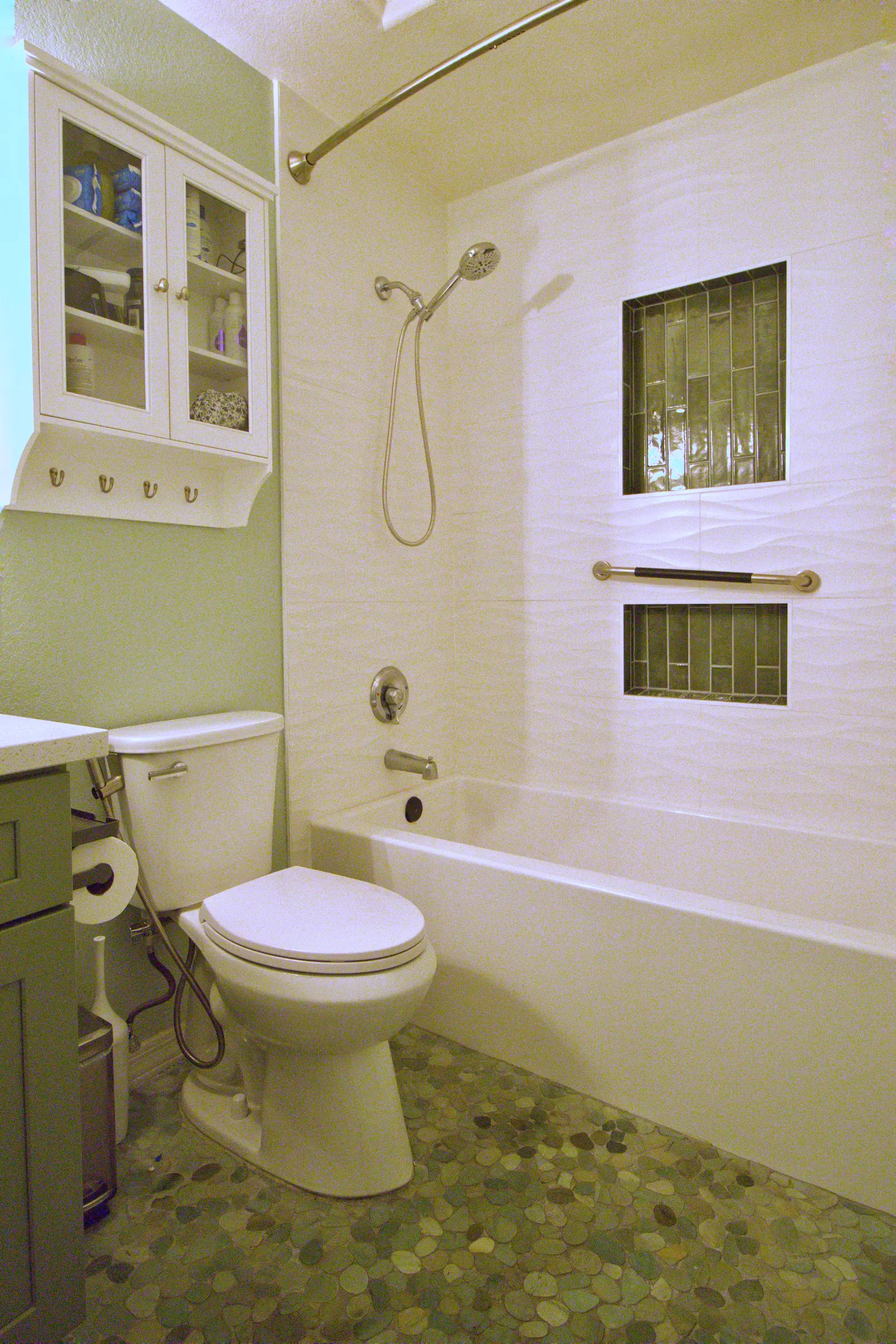 Guest bathroom in sage green with a pebble mosaic floor, a white tub with grab bar, glass mosaic shower niches and a wall cabinet