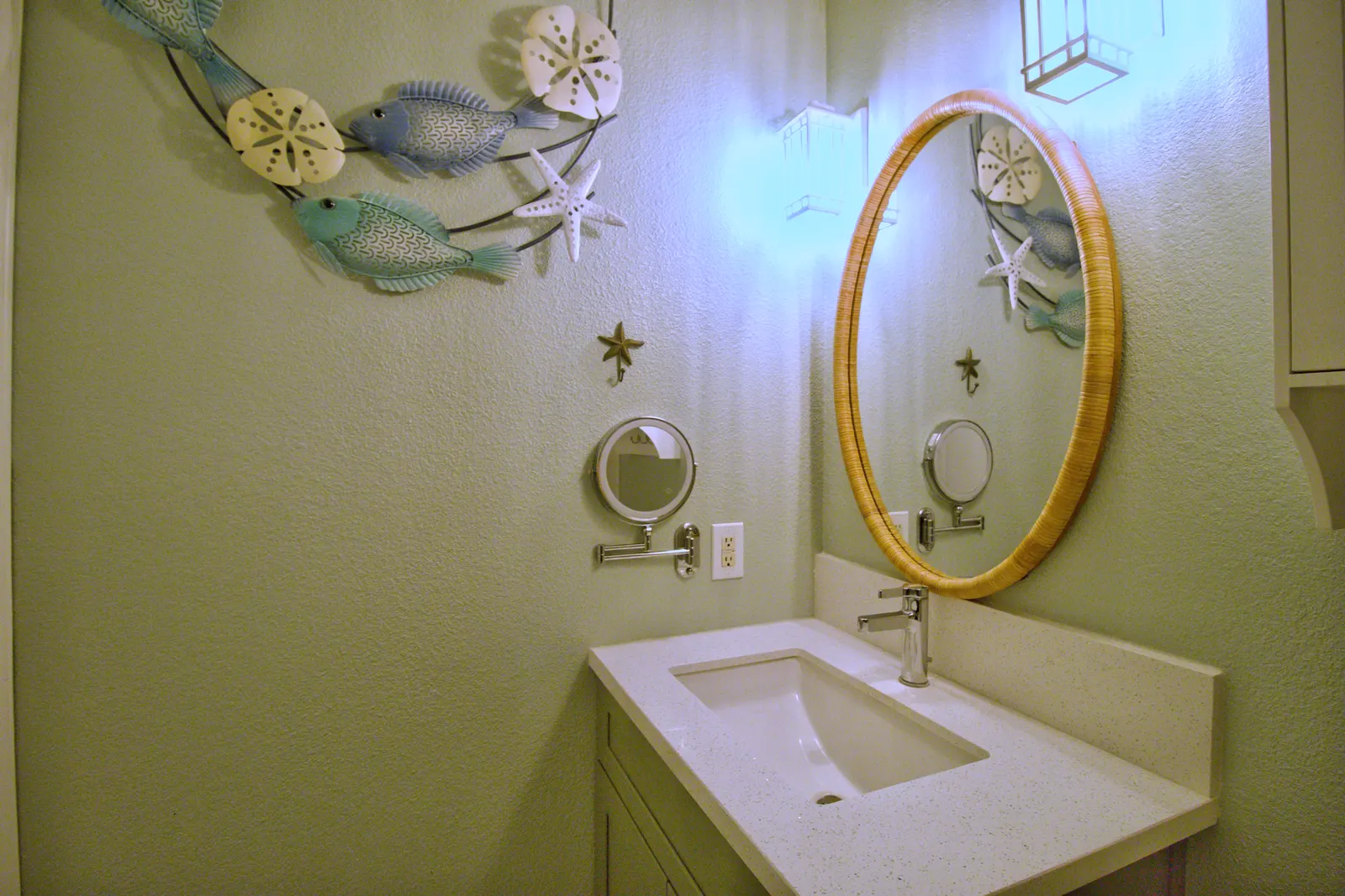 Oval rattan-framed mirror above a quartz vanity, with a metal wall sculpture of fish and sand dollars alongside