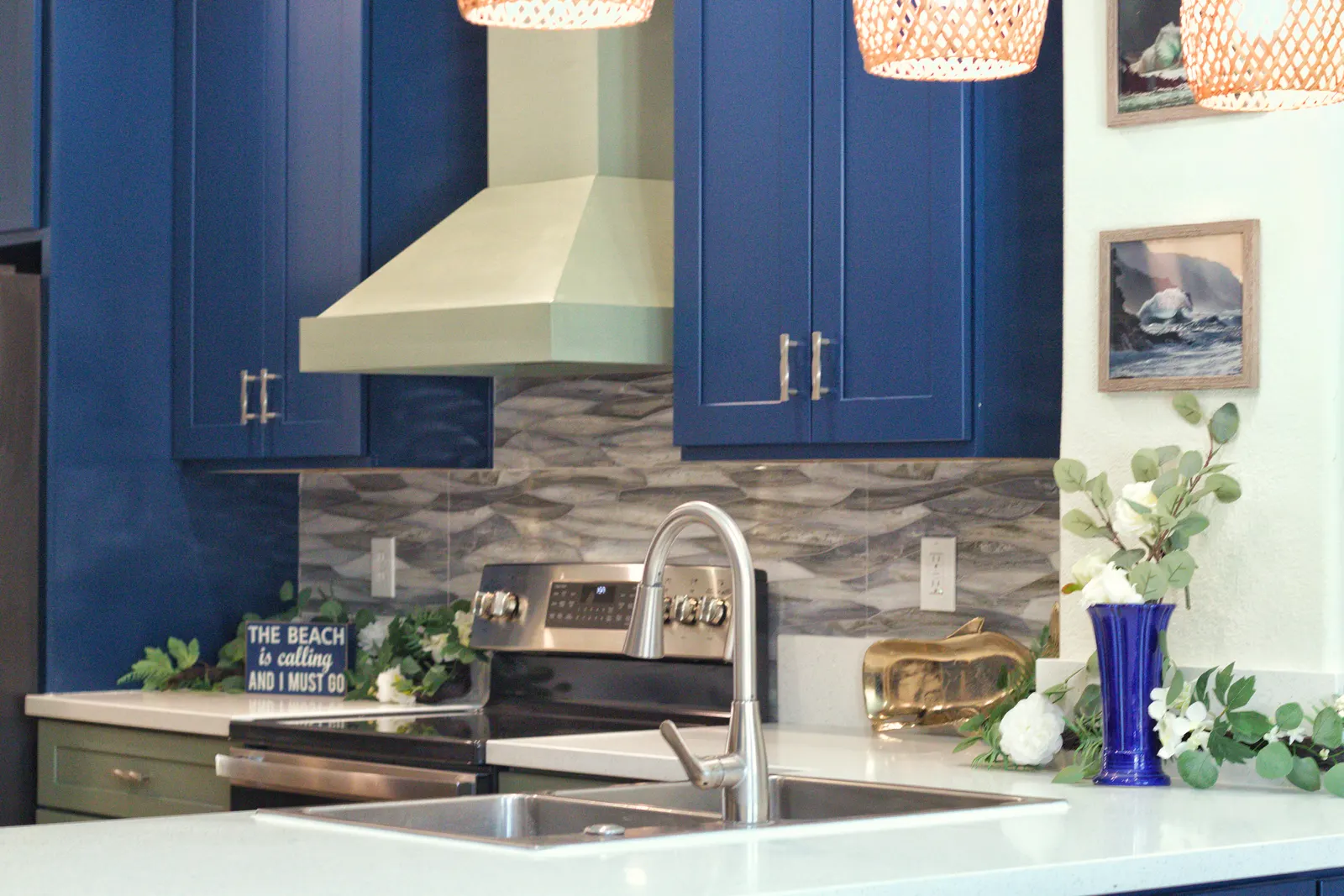 Sage range hood framed by navy cabinets, with woven rattan pendants and framed ocean prints alongside