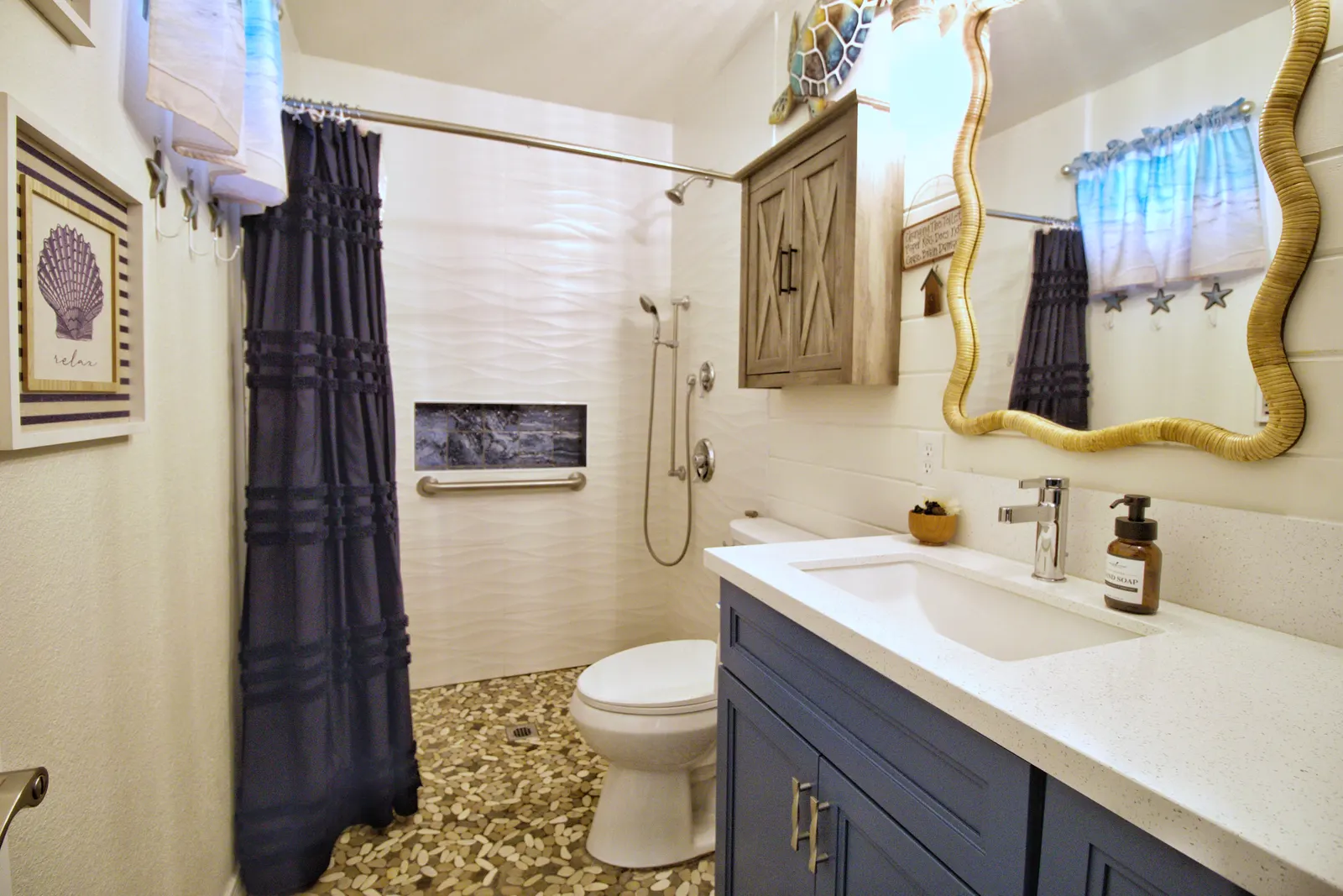 Master bathroom with a navy vanity under white quartz, a wavy rattan-framed mirror, a pebble tile floor and a grab bar in the shower
