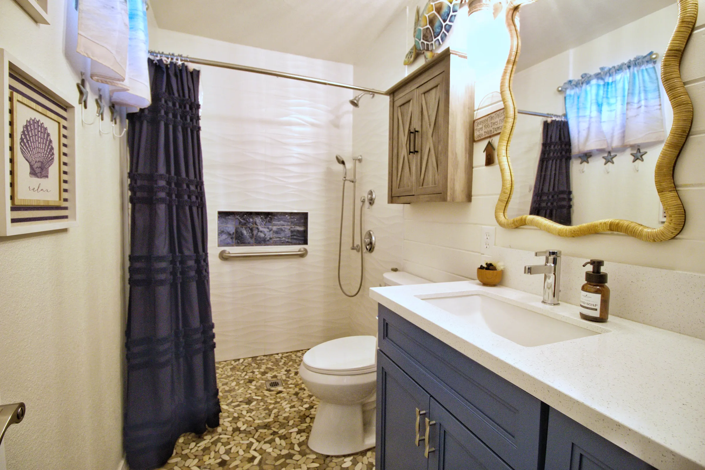 Master bathroom with a navy vanity under white quartz, a wavy rattan-framed mirror, a pebble tile floor and a grab bar in the shower