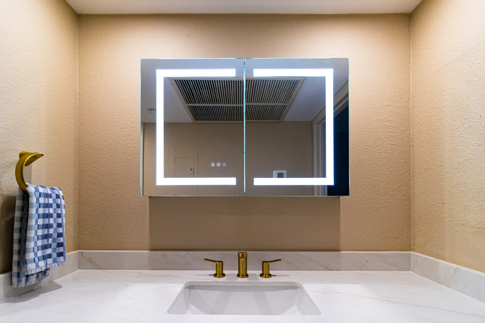 LED-backlit medicine cabinet mirror above a marble-look vanity top with a brass widespread faucet