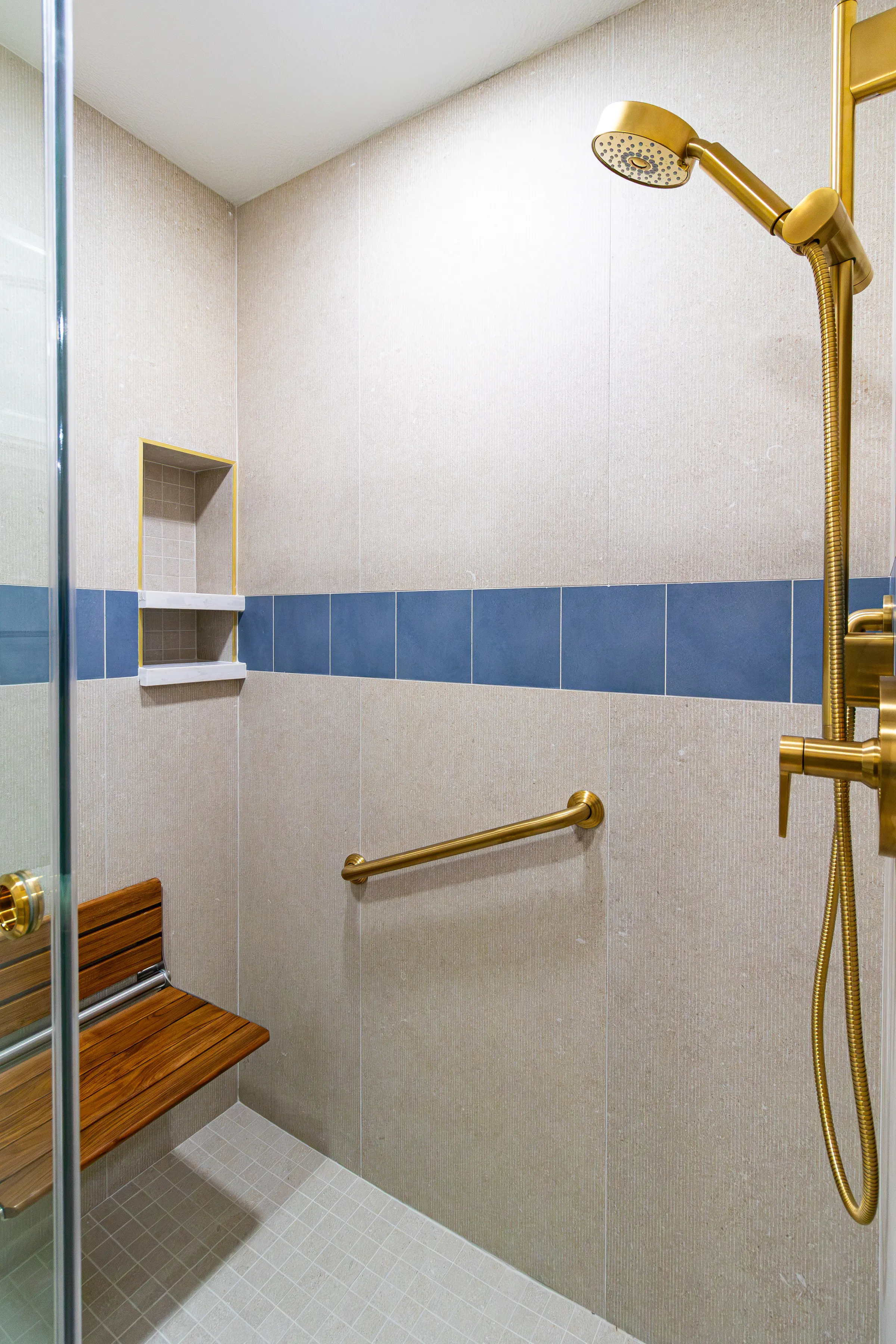 Curbless shower with a brass rain head and handheld sprayer, teak fold-down seat, brass grab bars, a tiled niche and a blue accent tile band