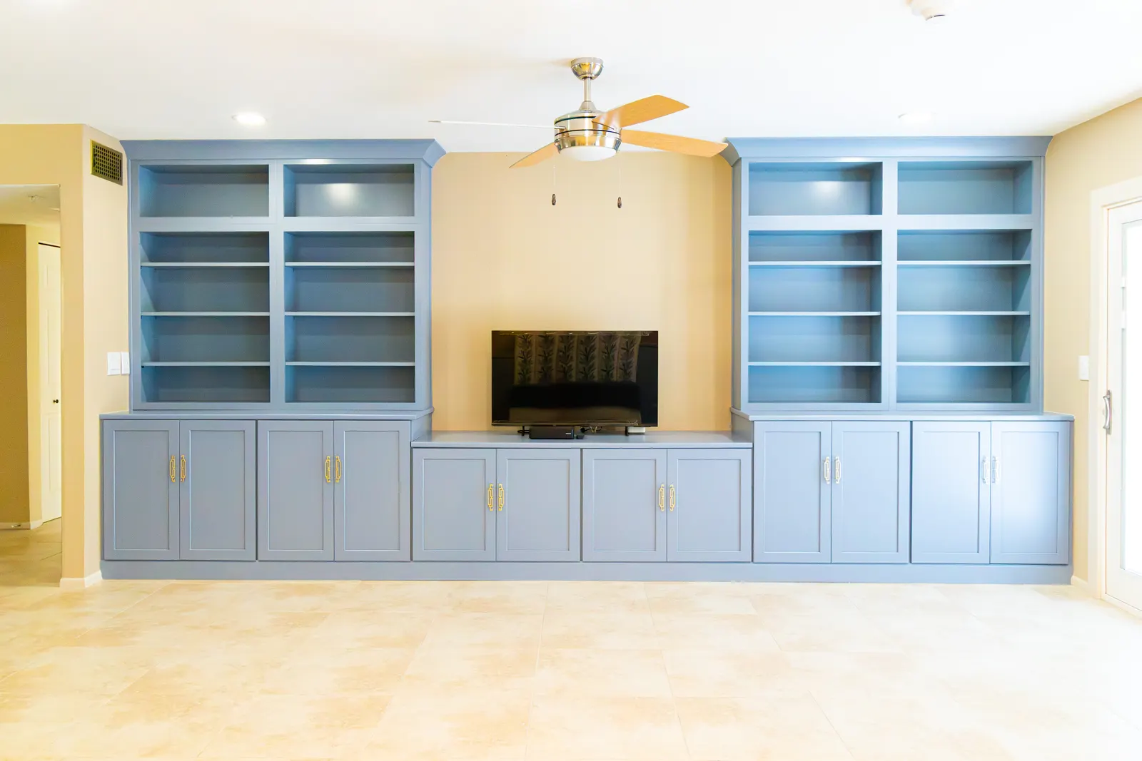 Full wall of blue built-in shelving and cabinets framing a television, with a ceiling fan and travertine tile floor