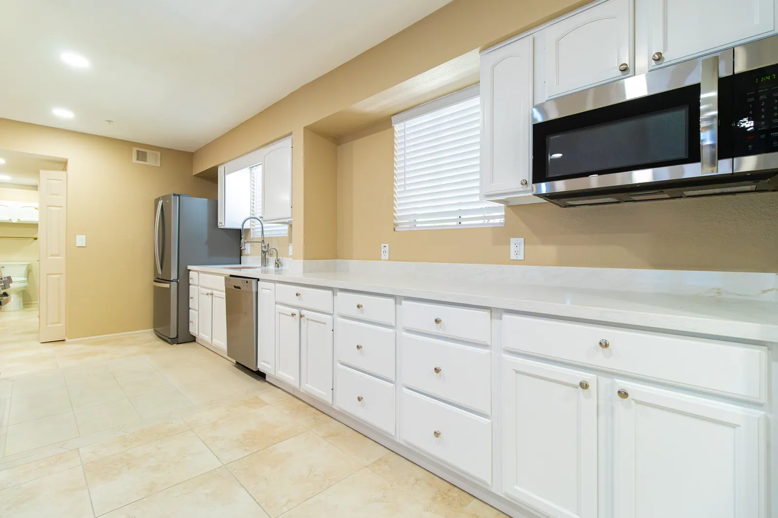 Long galley run of white cabinets under a quartz counter, with stainless refrigerator, dishwasher and over-range microwave